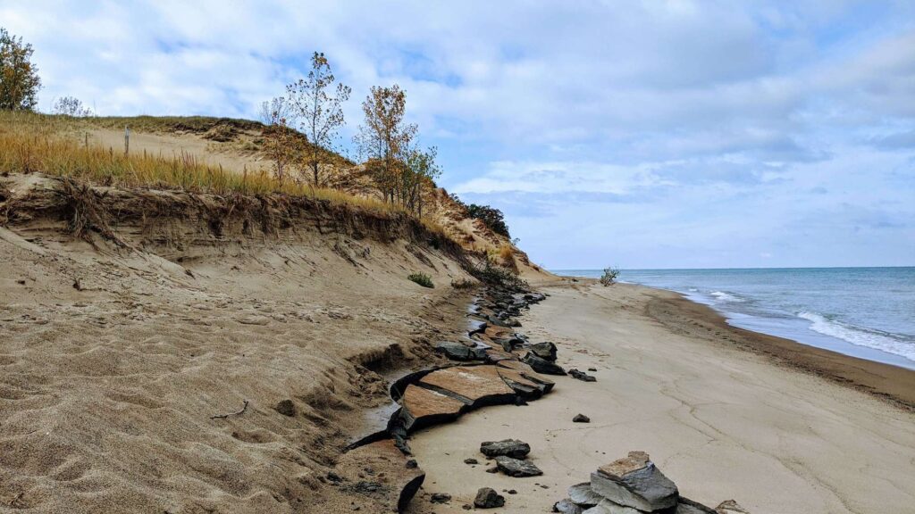 Large chunks of sheet rock crack and break off onto the beach.  The sky is cloudy and the water is bright blue.  Large sandy cliffs outline the left edge with trees and grass almost ready to fall into the ocean. 