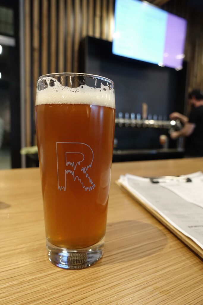A dark amber, cloudy beer sitting at the edge of the bar.  A menu is out of focus to the right, and a bartender pulls a glass of beer from the taps in the background.  A large "R" logo is on the glass facing the camera. 