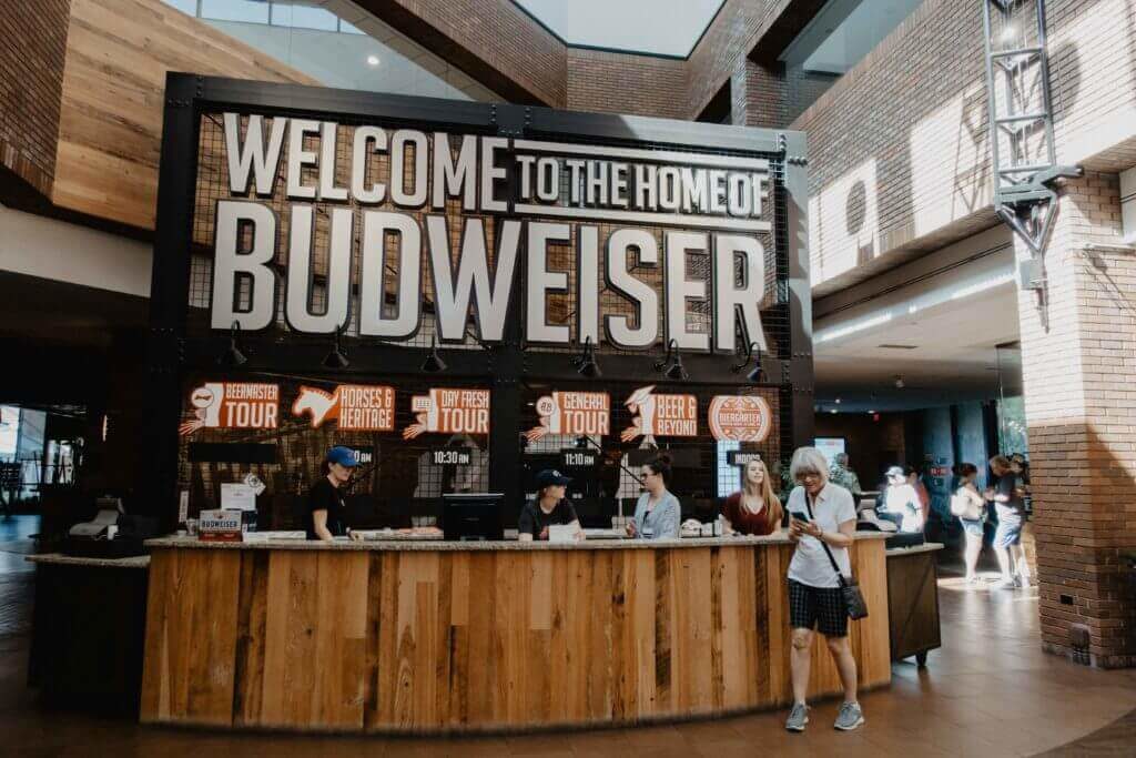 A large sign that reads "Welcome to the Home of Budweiser" in an open room.  Lots of people stand nearby, waiting in line for tours. 