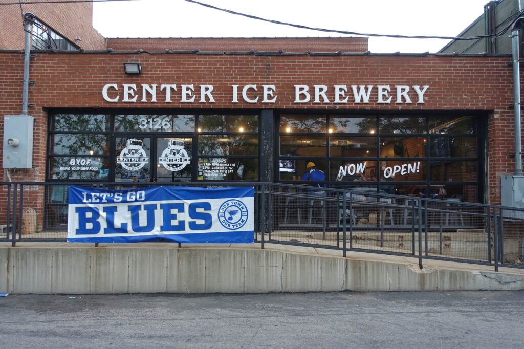 A Small brick building with a white sign that reads "Center Ice Brewery" and a flag on their banister that says "Let's Go Blues"
