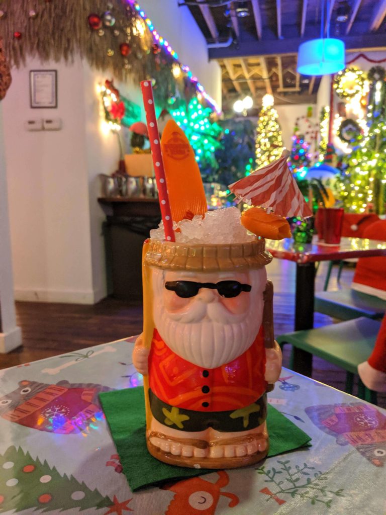 A large cup that is in the shape of Santa in a Hawaiian shirt. A small plastic surfboard garnish and a tiny drink umbrella poke out of the mound of crushed ice at the top.  Lots of christmas lights are out of focus in the background. 