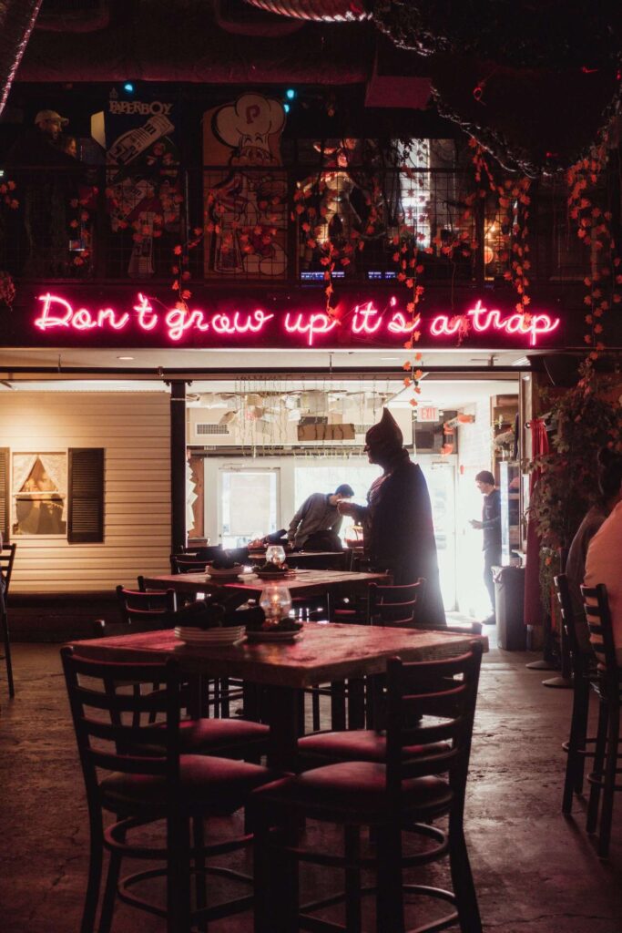 A neon sign in Nashville that says "Don't Grow up It's a trap" with a man dressed as Batman grabbing a drink right underneath the sign. 