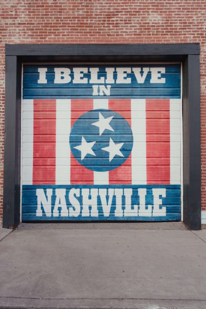 Mural in Nashville reading "I Believe in Nashville" painted by an image that resembles the Nashville City Flag. This mural is painted on a garage door in Nashville, but there are many other versions of this same mural across the city of Nashville.  Murals are hugely popular in Nashville, that's why this stop made the cut for our Nashville Weekend Itinerary