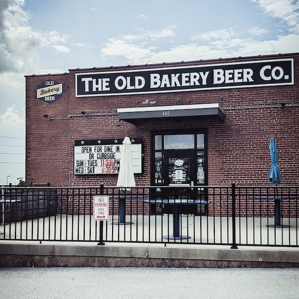 A small brick building in front of a blue sky with a black and white sign that says "Old Bakery Beer Co." There are a few umbrellas on their patio. 