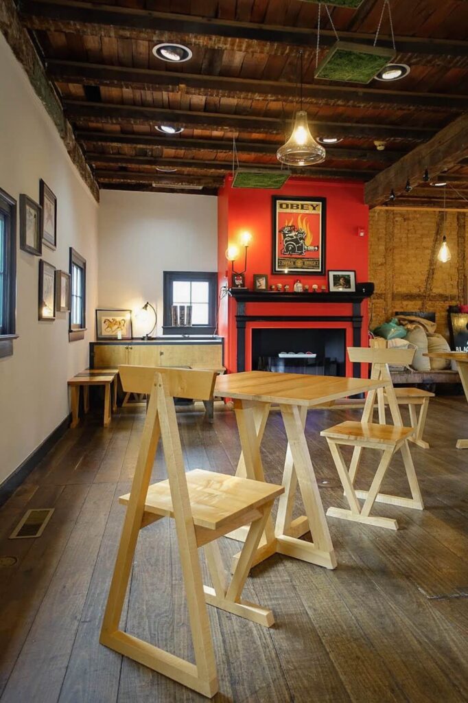 The inside of a small coffee shop.  The Chairs and tables are geometric and have odd angles.  They are light wood in contrast to all the dark wood of the floors and ceiling.  there is a bright red fireplace in the background. 