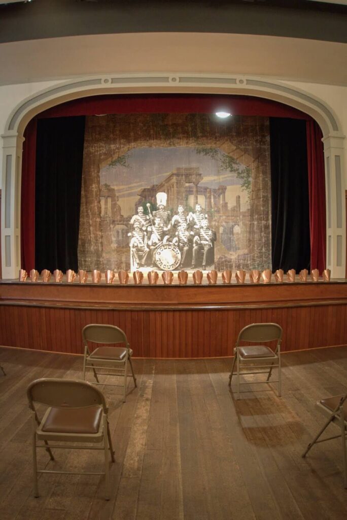 A small theater stage.  On the stage there is a cardboard cut out of a historic photo of a band that would have been on stage.  There are gorgeous brass seashell-shaped light fixtures lining the bottom of the stage.  Folding chairs fill the room. 