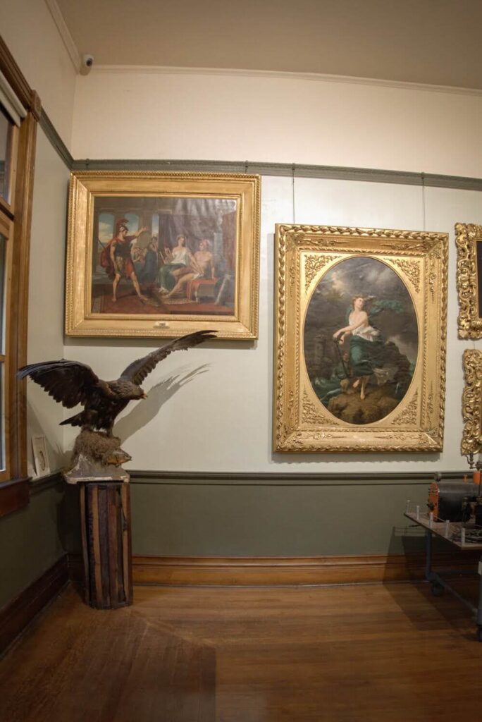 Many gold framed paintings hanging in a tiny museum in New Harmony.  There is also a large taxidermy golden eagle in the corner. 