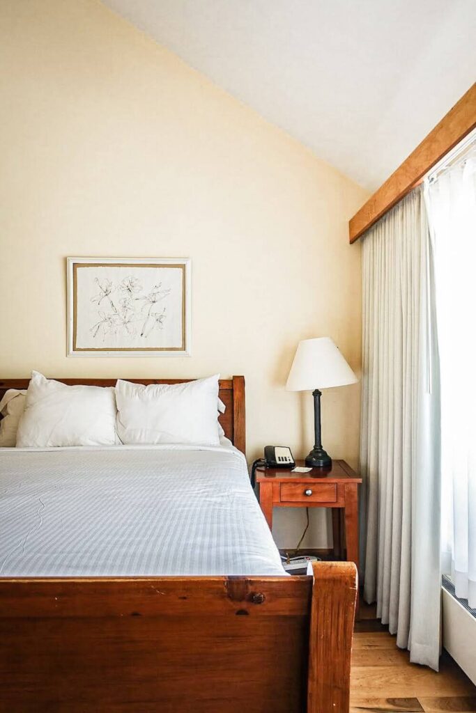Our bed at the New Harmony Inn.  It is a cherry oak bed frame contrasts bright white sheets on the bed.  The walls of the room are slightly beige.  A drawing of flowers hangs above the bed.  Everything is next to a wide open window shining a bright light into the room. 