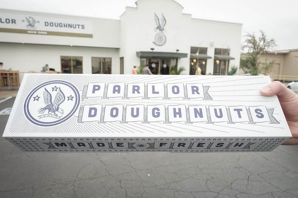The oblong donut box from Parlor Doughnuts is longer than it is wide. It is approximately 18 inches long, but only about 6 inches wide, making for a funny but memorable donut box shape.  It is held in one hand with the storefront out of focus in the background. 