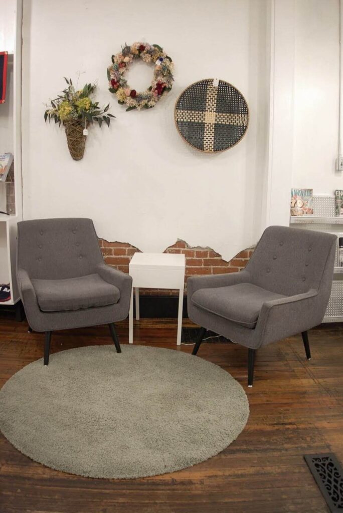 Two grey sofa chairs sit facing each other.  There is a white coffee table between them.  The wall is decorated with woven baskets and floral arrangements that are for sale. 