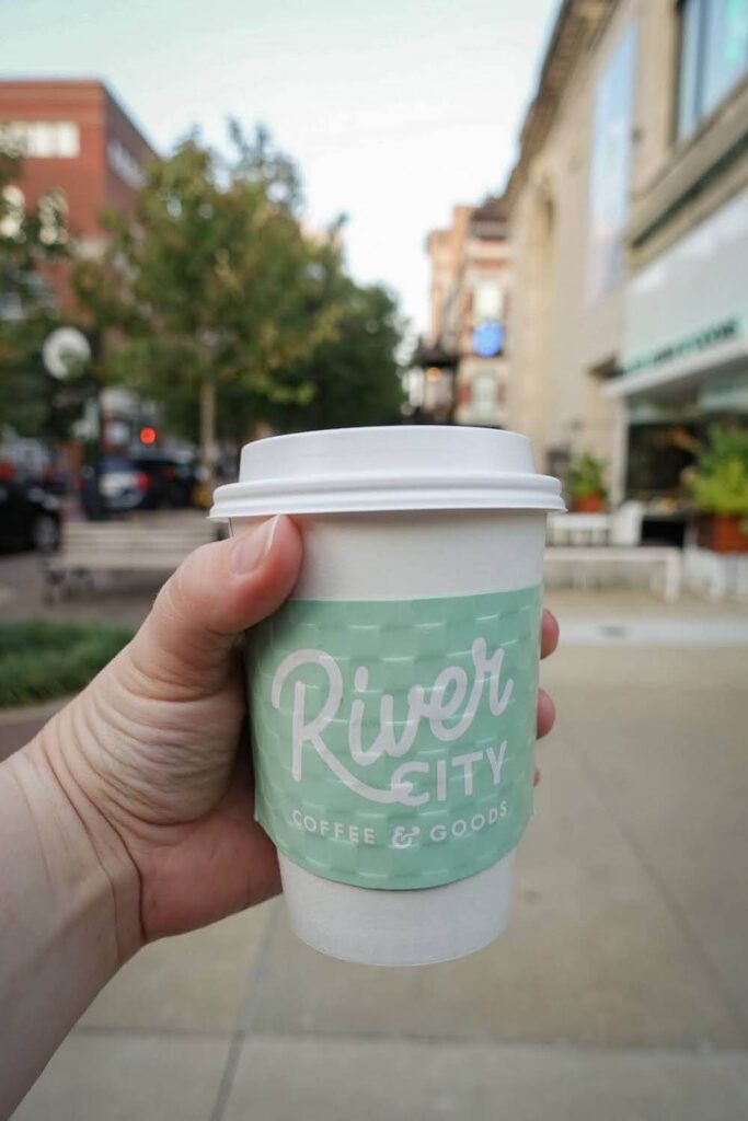 A simple white coffee cup with a light green sleeve that reads "River City Coffee & Goods." The streets of downtown Evansville are out of focus in the background. 