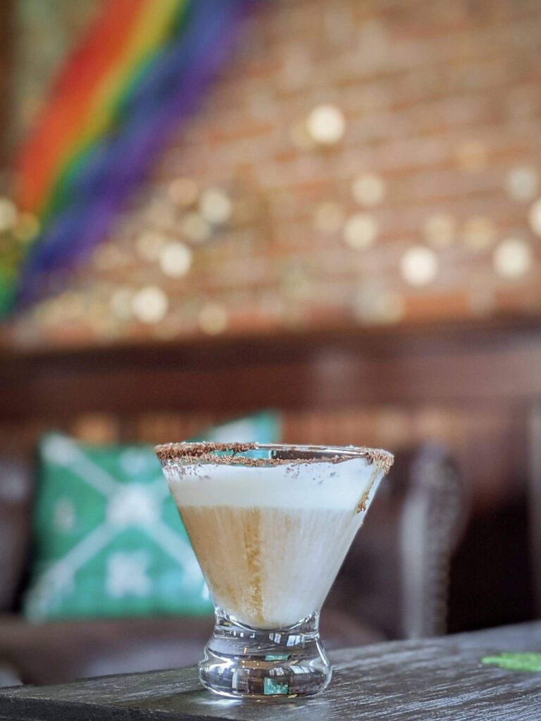 A creamy cocktail drink with a chocolate rim served in a stemless martini glass.  In the background there is a rainbow and pieces of gold on the wall. 