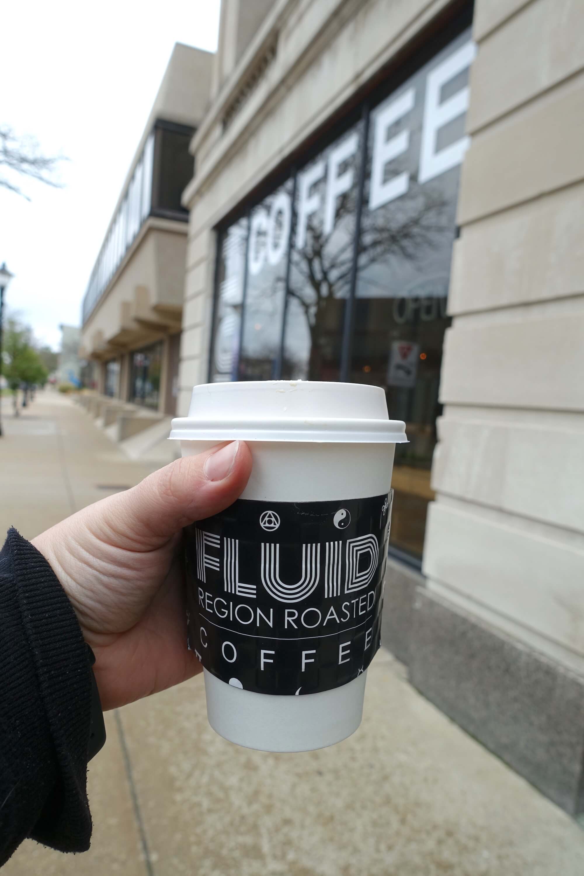 A coffee cup held up in front of a white building with the word COFFEE out of focus in the window. The sleeve of the coffee cup says Fluid Region Roasted Coffee - Best things to do near Indiana Dunes National Park